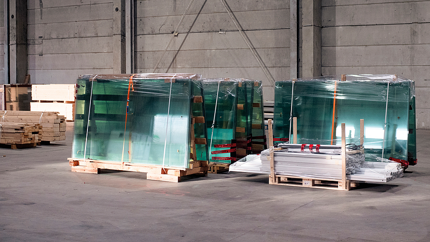 Mehrere große Glasscheiben, verpackt und auf Holzpaletten gelagert, stehen in einer Industriehalle.