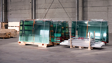 Mehrere große Glasscheiben, verpackt und auf Holzpaletten gelagert, stehen in einer Industriehalle.