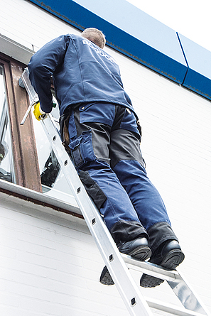 Ein Mann steht mit auf einer Leiter, die an einer Häuserwand lehnt und bis zu einem Fenster reicht, und arbeitet am Fenster.