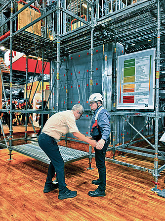 Zwei Männer in einer Messehalle stehen an einem Gerüst; einer befestigt ein temporäres Lifeline-System zur Absturzsicherung am anderen.