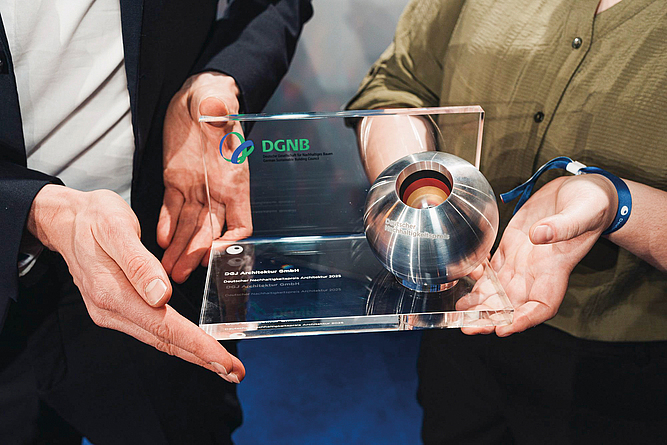 Zwei Personen halten gemeinsam eine Auszeichnung der DGNB mit einer silbernen Kugel in einem transparenten Rahmen.