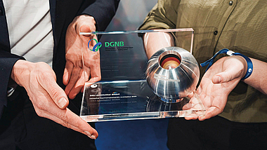 Zwei Personen halten gemeinsam eine Auszeichnung der DGNB mit einer silbernen Kugel in einem transparenten Rahmen.