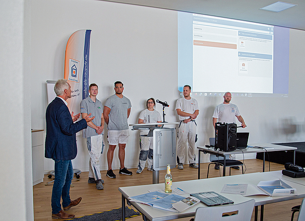 Fünf Auzubildende in Arbeitskleidung und ein älterer Mann links daneben präsentieren etwas vor einer Leinwand.