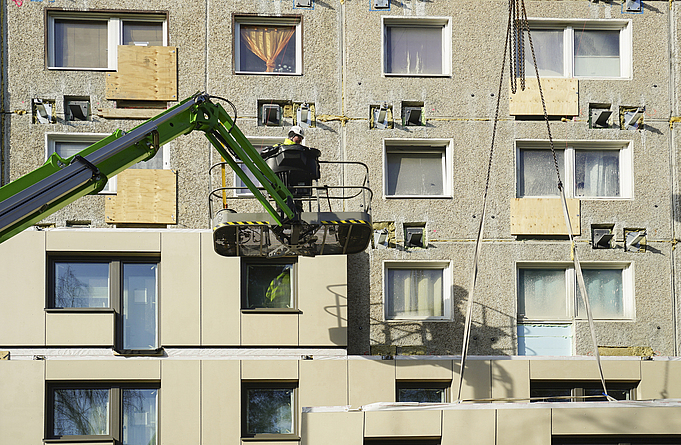 Person mit Schutzkleidung und Helm auf einer Arbeitsbühne vor der halbsanierten Fassade des Plattenbaus.
