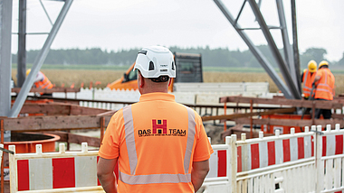 Bauarbeiter in Warnkleidung und Helm steht vor einer Baustelle mit Absperrung und Stahlkonstruktion.