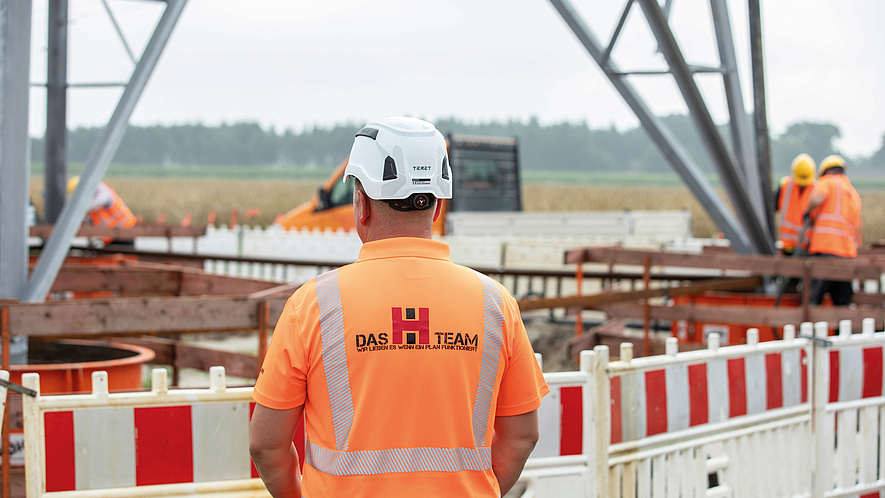 Bauarbeiter in Warnkleidung und Helm steht vor einer Baustelle mit Absperrung und Stahlkonstruktion.