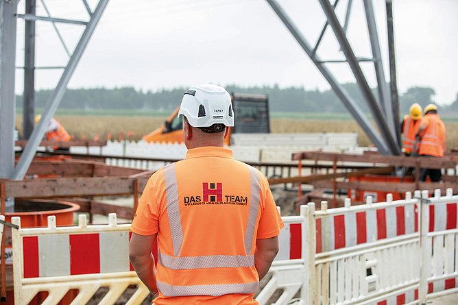 Bauarbeiter in Warnkleidung und Helm steht vor einer Baustelle mit Absperrung und Stahlkonstruktion.