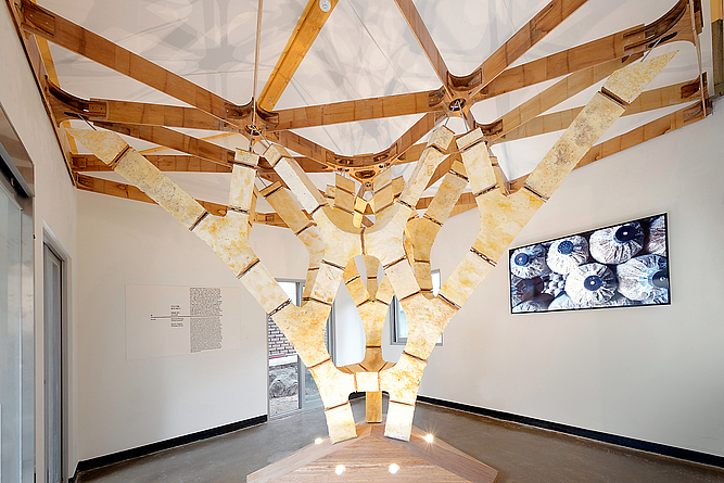 Ausstellungsraum, in dessen Zentrum ein verzweigter, künstlicher Baum aus hellen Materialien steht. Der Baum trägt die Decke des Raumes.
