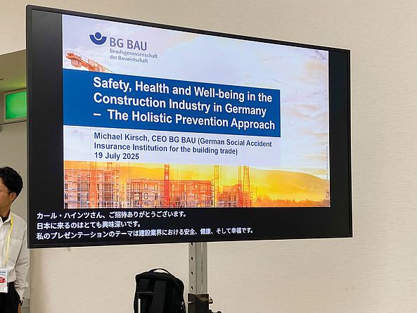 Präsentationsfolie auf einem Bildschirm mit dem BG BAU-Logo und dem Titel „Safety, Health and Well-being in the Construction Industry in Germany – The Holistic Prevention Approach“.