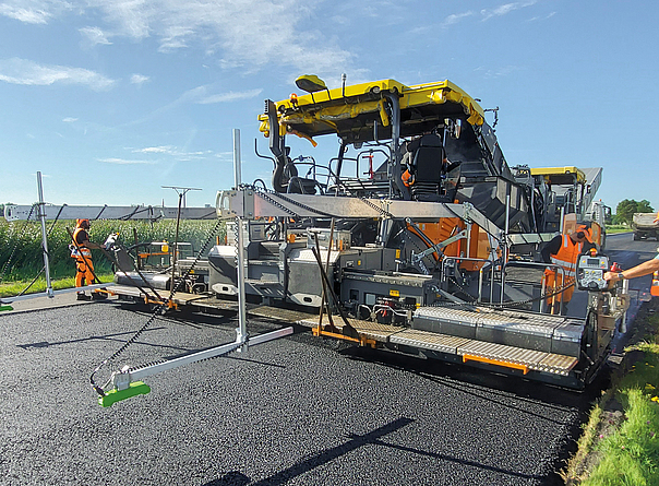 Arbeitsplatzmessung beim Straßenbau: Eine Straßenbaumaschine trägt Asphalt auf, während Messgeräte und Beschäftigte den Arbeitsprozess überwachen.