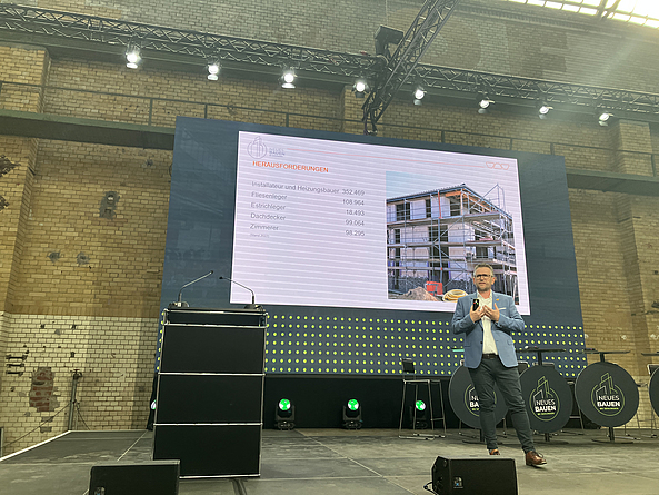 Jens Göke, Schlüter Systems, startete mit den Herausforderungen in der Branche und stellte dann Systemlösungen für den effizienten Neu- und Bestandsbau vor. Jens Göke auf der Bühne, hinter ihm eine Präsentationswand mit einer Folie.