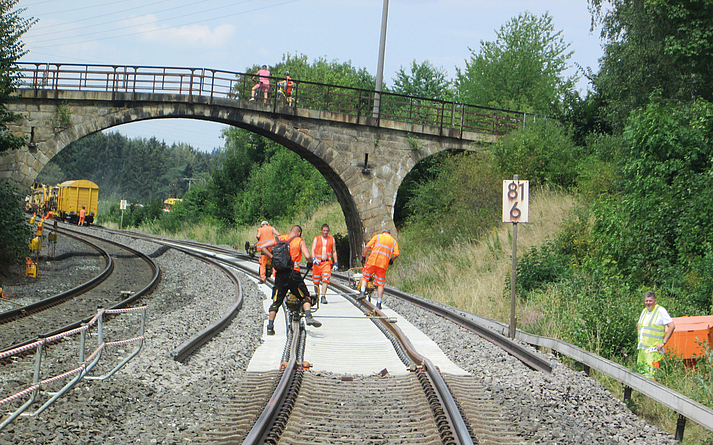 Mehrere Personen in Warnkleidung arbeiten an einer zweigleisigen Bahnstrecke. Im Hintergrund überspannt eine Steinbrücke die Gleise.