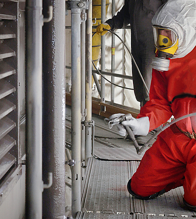 Eine Person mit rotem Schutzanzug und Atemschutz strahlt auf ein Element der Fassade.