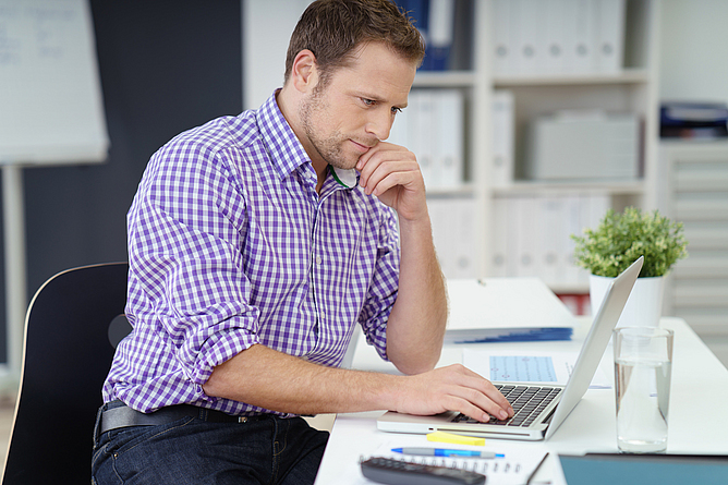 Ein Mann sitzt an einem Schreibtisch. Er arbeitet am Laptop.