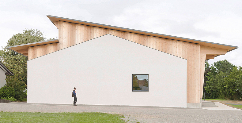 Modernes Gebäude mit heller Fassade und Holzelementen, davor eine Person auf einer Grünfläche.