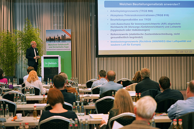 Blick in den Vortragsraum. Die Teilnehmenden sitzen an Tischen und sind von hinten zu sehen. Vorne referiert ein Mann am Stehpult.