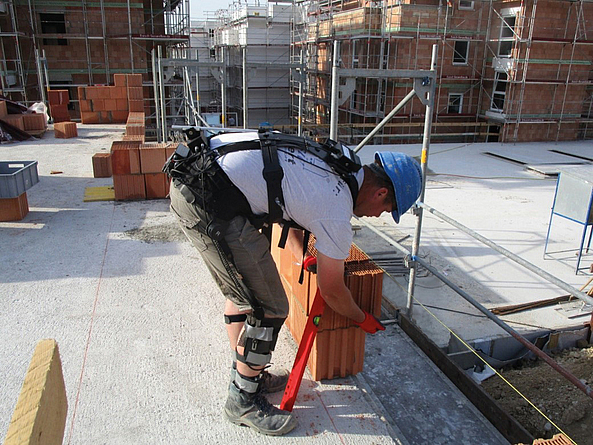 Auf einer Baustelle hebt ein Bauarbeiter mit Schutzhelm Mauersteine an. Er trägt zur Messung der ergonomischen Belastung ein Messsystem am Oberkörper. 