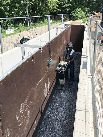 Eine Person mit Helm, Gehörschutz und Atemschutz arbeitet mit einer Verdichtungsmaschine auf einem schmalen, mit Schotter bedeckten Abschnitt. Messgeräte sind am Rand des Grabens angebracht.