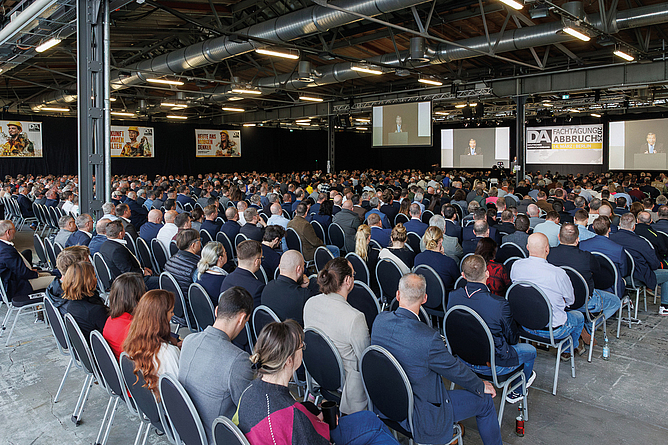 Große Anzahl von Teilnehmenden sitzt in einem Veranstaltungssaal und verfolgt eine Präsentation auf mehreren Leinwänden.
