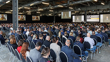 Große Anzahl von Teilnehmenden sitzt in einem Veranstaltungssaal und verfolgt eine Präsentation auf mehreren Leinwänden.