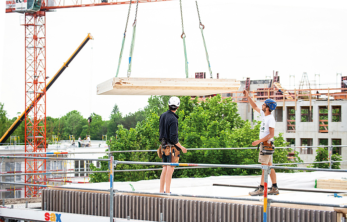 Zwei Männer mit Helmen stehen auf einer gesicherten Dachfläche und nehmen ein Holzbauteil entgegen, welches von einem Kran herabgesenkt wird.