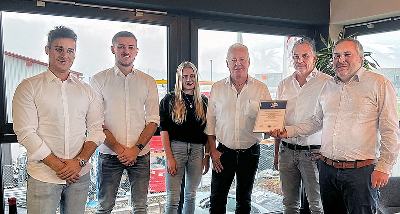 Fünf Männer in weißen Hemden und eine Frau mit schwarzem Oberteil stehen vor Glasfront.