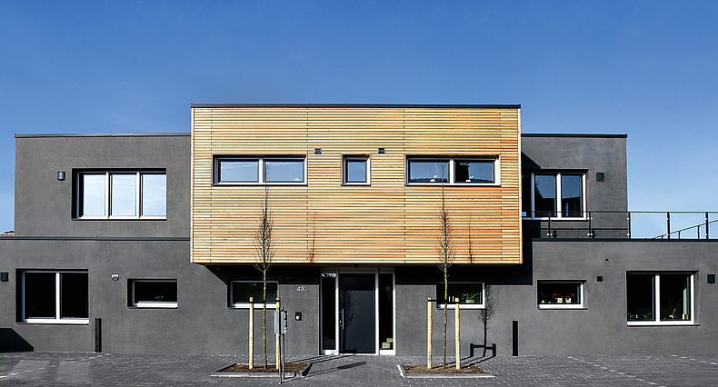 Frontalblick auf graues Betonhaus mit einem Vorbau aus Holzpaneelen. 
