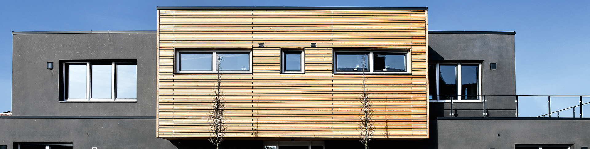 Frontalblick auf graues Betonhaus mit einem Vorbau aus Holzpaneelen. 
