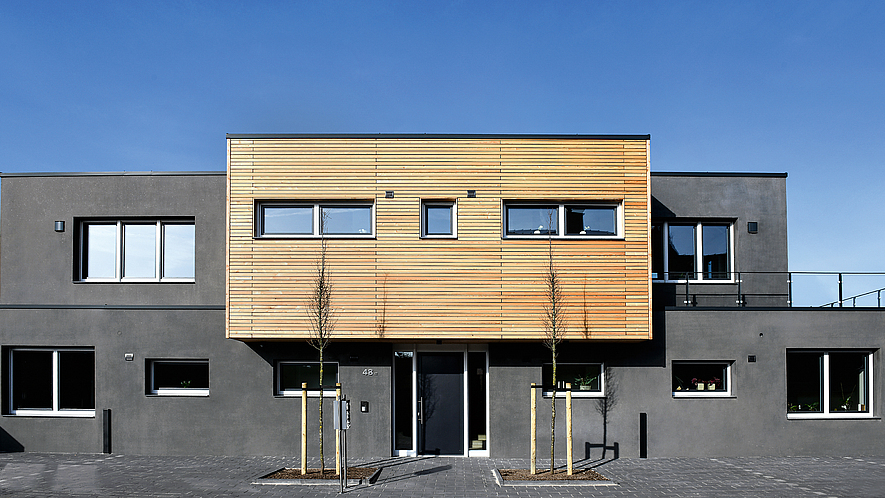 Frontalblick auf graues Betonhaus mit einem Vorbau aus Holzpaneelen. 