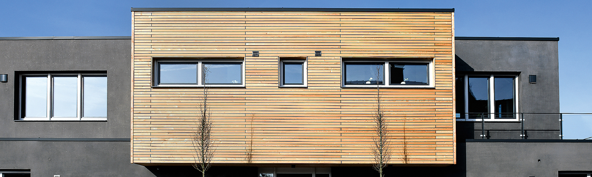 Frontalblick auf graues Betonhaus mit einem Vorbau aus Holzpaneelen. 