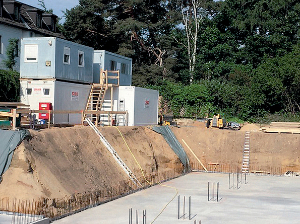 Die Auflast durch Container am Böschungsrand ist zu hoch und deshalb nicht zulässig. Aufeinander gestapelte Container am Böschungsrand einer großen Baugrube, in welche Leitern führen und in deren Grund Bewehrungseisen herausragen.