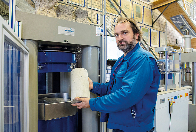 Ingo Fenneker macht im Bautechnischen Zentrallabor Belastungstests Ein Mann steht vor einer geöffneten Messanlage und hält einen Betonklotz in der Hand.