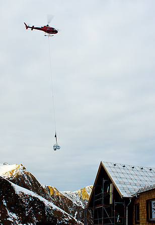 An einem Hubschrauber in der Luft hängt ein Seil mit Materialien. Im Vordergrund ist der obere Teil der Hütte abgebildet.