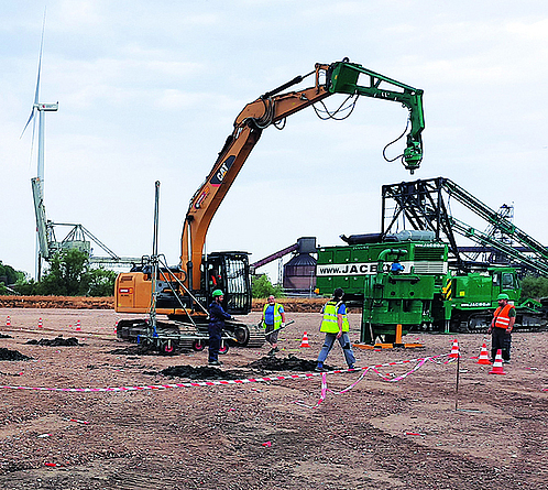 Auf einer Sandfläche sind in der Ferne zahlreiche Bohrlöcher zu sehen. Daneben befinden sich ein Bagger und drei Personen in Schutzkleidung