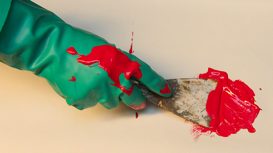 Ein Nahaufnahme-Foto zeigt eine mit einem grünen Gummihandschuh bekleidete Hand, die mit einem Spachtel leuchtend rote Farbe auf einer hellen Oberfläche verteilt.