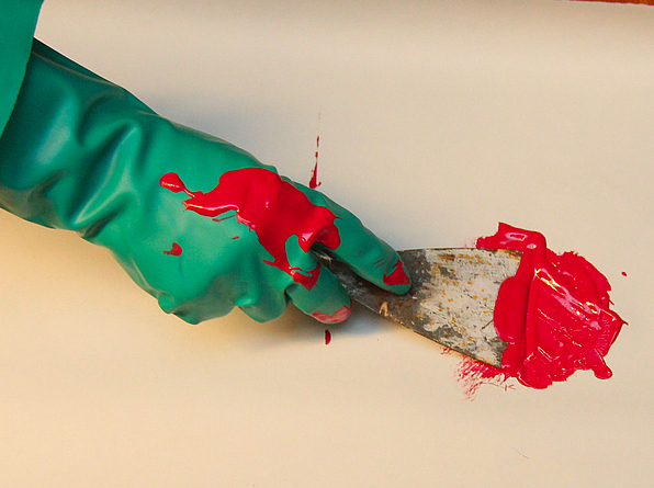 Ein Nahaufnahme-Foto zeigt eine mit einem grünen Gummihandschuh bekleidete Hand, die mit einem Spachtel leuchtend rote Farbe auf einer hellen Oberfläche verteilt.
