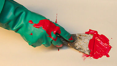 Ein Nahaufnahme-Foto zeigt eine mit einem grünen Gummihandschuh bekleidete Hand, die mit einem Spachtel leuchtend rote Farbe auf einer hellen Oberfläche verteilt.