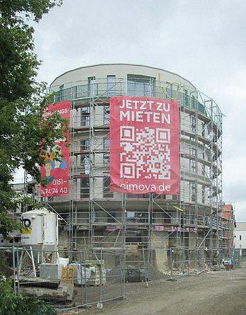 Rundes Wohngebäude, das eingerüstet ist. Im angrenzenden, mit einem Zaun abgetrennten Bereich befinden sich Baumaterialien und Baugeräte. 