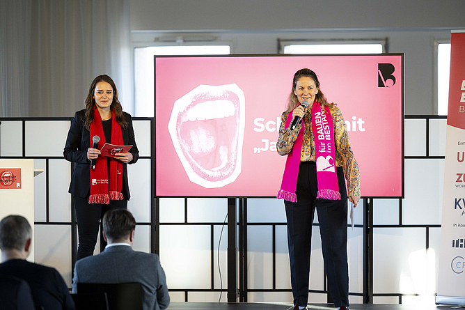 Zwei Frauen stehen vor auf einem Podium und sprechen zum Publikum.