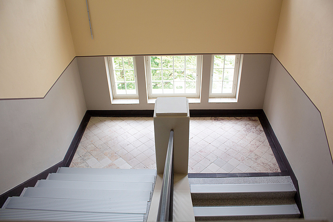 Blick nach unten in das Treppenhaus, sodass die Treppen mit Stufenbelag, die Wandgestaltung des Treppenflurs und die Terazzofliesen zu sehen sind.