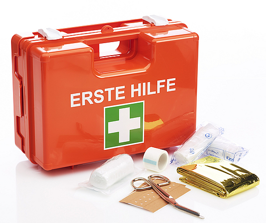 Roter Koffer mit weißer Schrift "Erste Hilfe" und darunter das weiß-grüne Symbol als Kreuz. Vor dem Koffer liegen Verbandsmaterial und eine Schere aus.