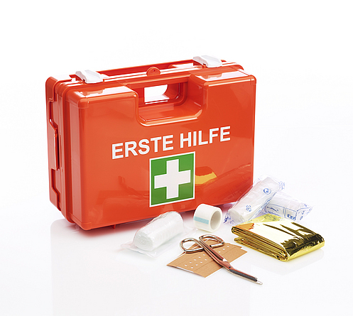 Roter Koffer mit weißer Schrift "Erste Hilfe" und  darunter das weiß-grüne Symbol als Kreuz. Vor dem Koffer liegen Verbandsmaterial und eine Schere aus.