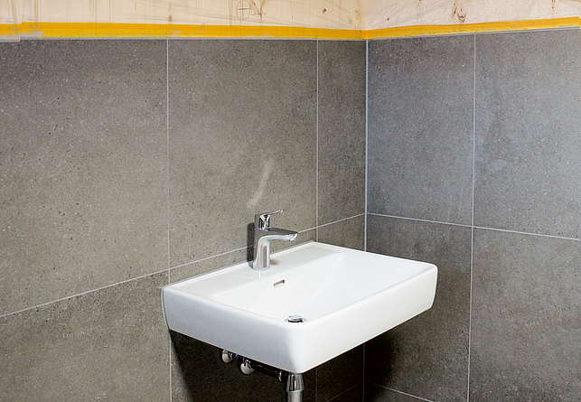 Waschbecken mit Armatur, das sich in der Ecke eines grau gefliesten Sanitärraums befindet.