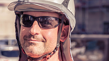 Portrait eines Bauarbeiters mit Sonnenbrille, Schutzhelm mit Vier-Punkt-Kinnriemen über dem ein Nackenschutz mit Sonnenblende gezogen ist. 