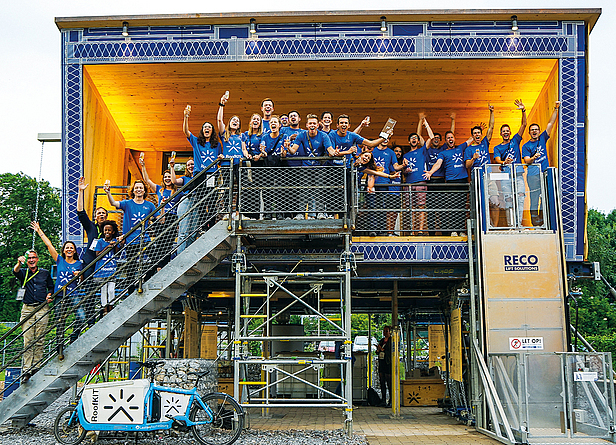 Das Gewinner-Projekt: „RoofKIT“, entwickelt von Studierenden des Karlsruher Instituts für Technologie (KIT) und Prof. Dirk E. Hebel Ca. 40 Menschen im blauen T-Shirt jubeln im ersten Stock auf der Treppe des aufgestockten Gebäudes.