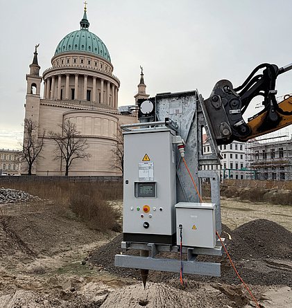 Bohrlochstopfmaschine wird mit einem Bagger auf einer Baustelle in den Boden eingesetzt, im Hintergrund steht ein Kuppelbau.