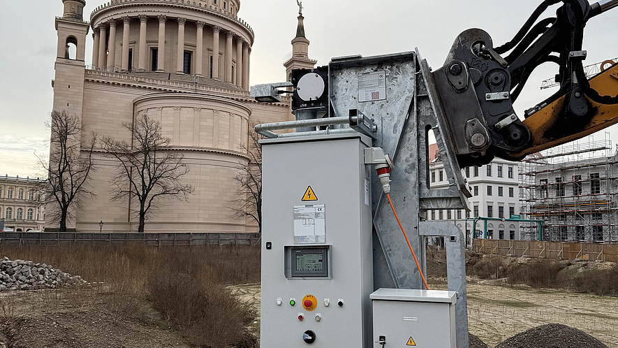 Bohrlochstopfmaschine wird mit einem Bagger auf einer Baustelle in den Boden eingesetzt, im Hintergrund steht ein Kuppelbau.