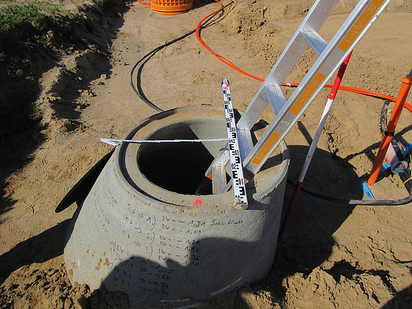 Offener Betonschacht auf einer Baustelle mit angelegter Leiter und Messlatte.