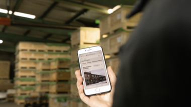 Smartphone im Lager Person hält ein Smartphone in der Hand, auf dem eine App läuft. Im Hintergrund sind Paletten zu sehen.