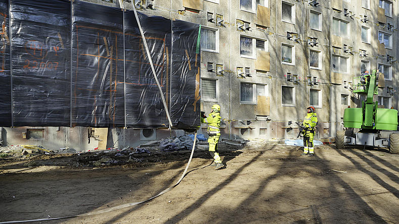 Ein verpacktes Fassadenmodul wird mit einem Kran Richtung Boden gesetzt. Eine Person begleitet das Aufsetzen am Führungsseil. Eine andere Person schaut zu.
